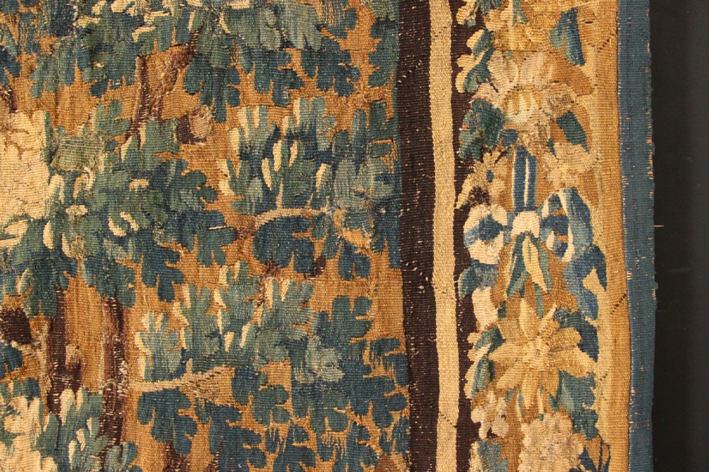 A large 18th-century hand-woven verdure tapestry depicting a dense woodland scene with foliage, animals, and distant architecture within a decorative floral border. View X