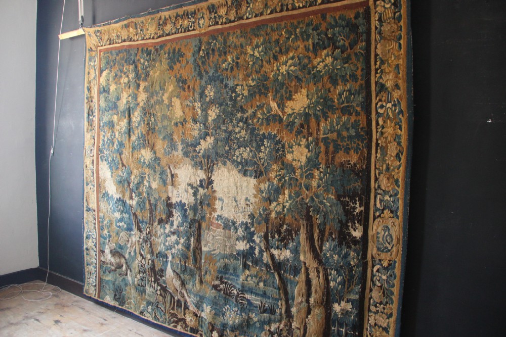 A large 18th-century hand-woven verdure tapestry depicting a dense woodland scene with foliage, animals, and distant architecture within a decorative floral border. View XI