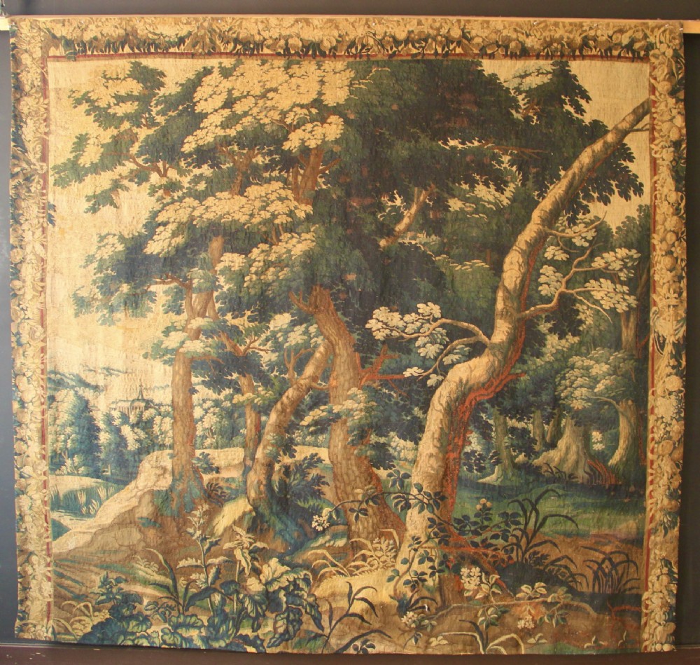 An 18th century verdure tapestry depicting a dense woodland scene with mature trees, foliage, and a distant landscape in muted greens and browns.