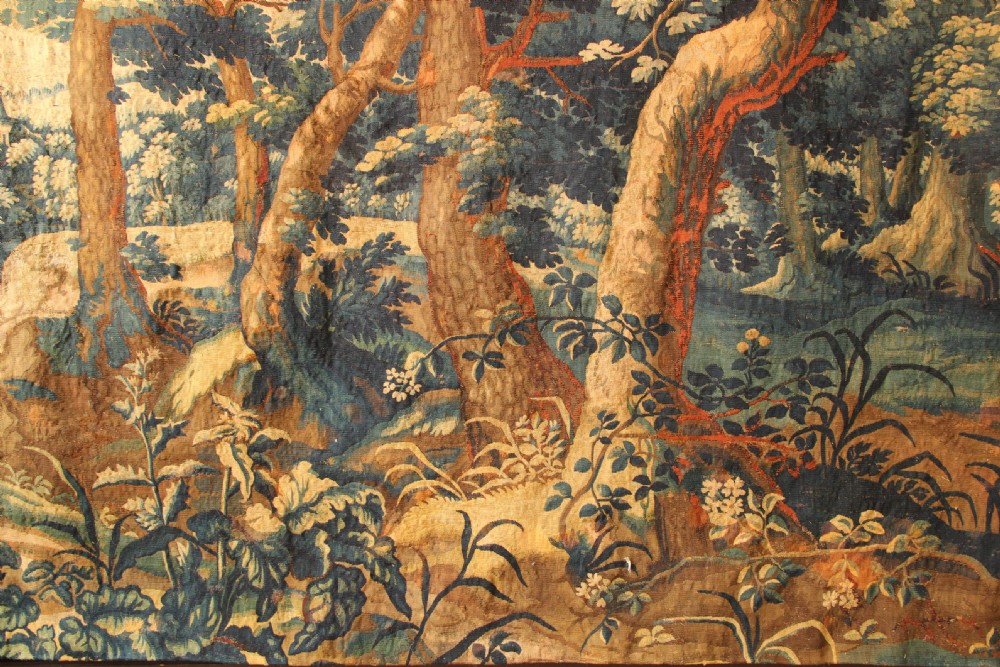 An 18th century verdure tapestry depicting a dense woodland scene with mature trees, foliage, and a distant landscape in muted greens and browns. View III