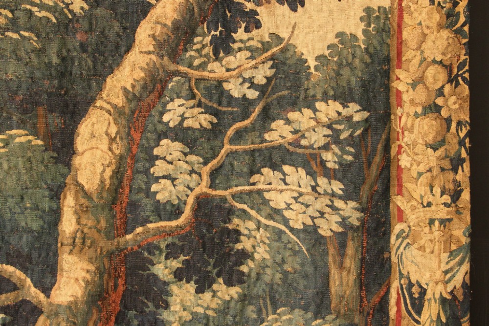 An 18th century verdure tapestry depicting a dense woodland scene with mature trees, foliage, and a distant landscape in muted greens and browns. View V
