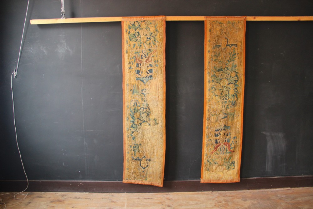 A pair of long vertical 17th-century woven tapestry borders featuring botanical foliage, urns, and floral motifs in yellow, green, and blue tones.