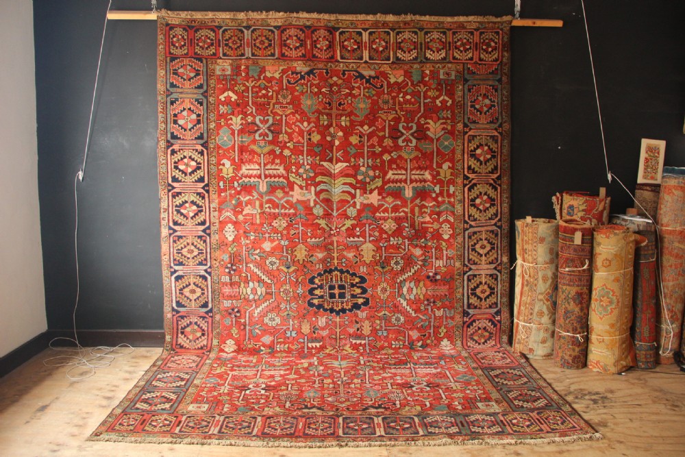 A large hand-knotted antique Heriz carpet with a rich terracotta red field, geometric floral motifs, and a navy blue central medallion.