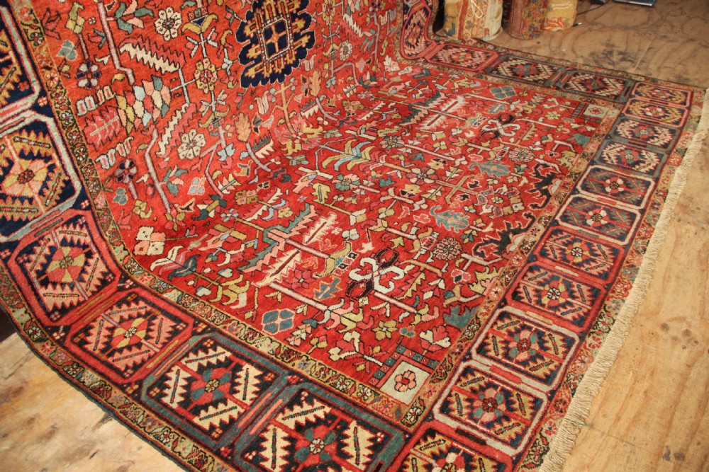 A large hand-knotted antique Heriz carpet with a rich terracotta red field, geometric floral motifs, and a navy blue central medallion. View X