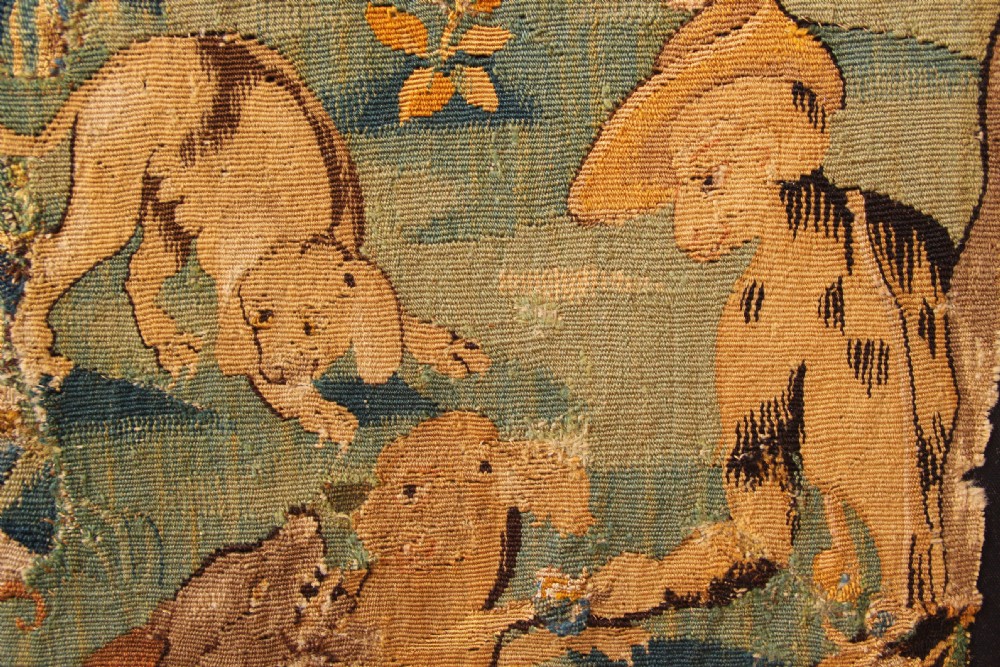 A seventeenth-century Flemish woven wool tapestry fragment depicting hounds in a wooded parkland landscape, mounted on a dark board. View II