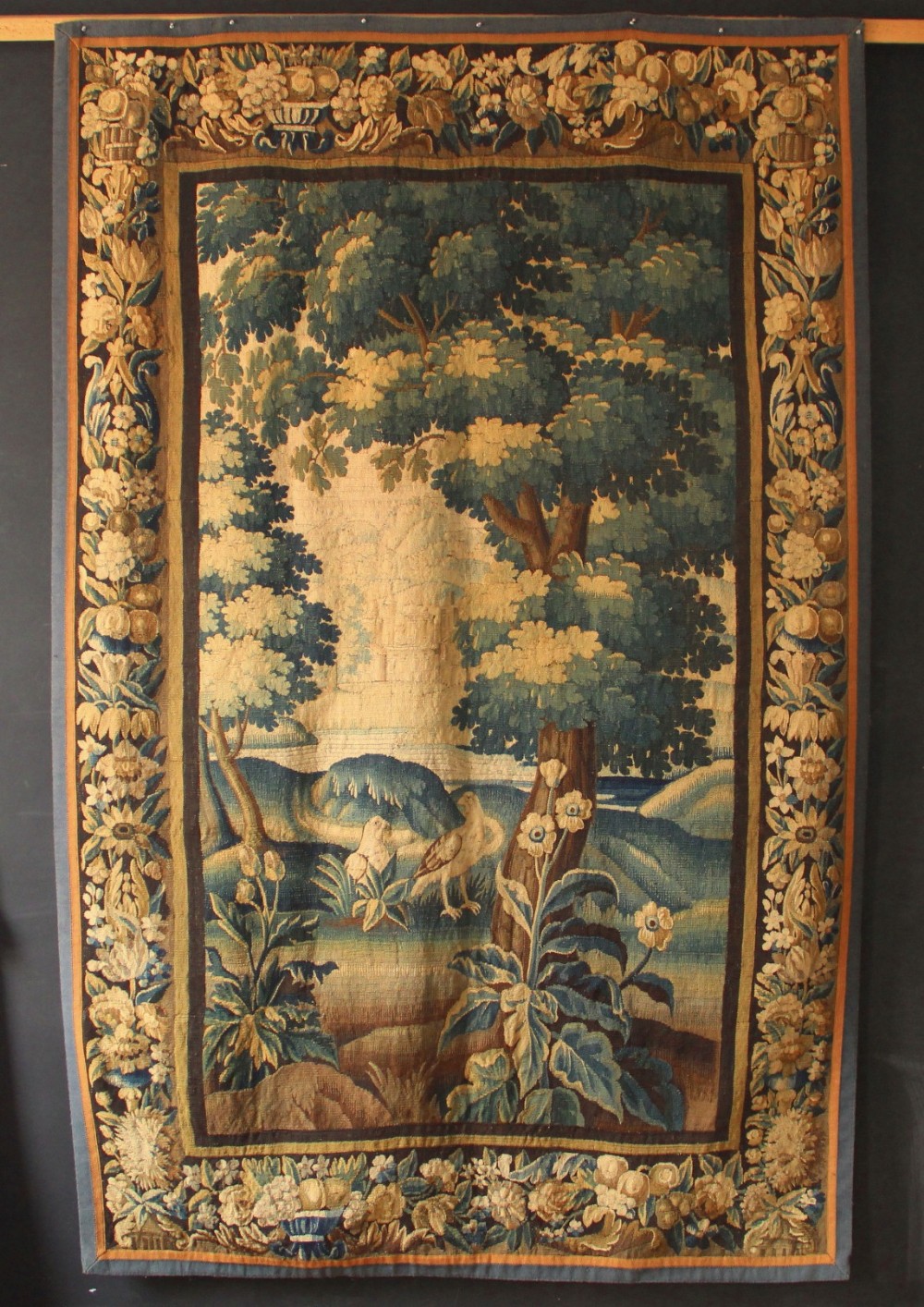 An 18th-century hand-woven Flemish verdure tapestry depicting a dense woodland scene with birds and a floral border in shades of green, cream, and brown.