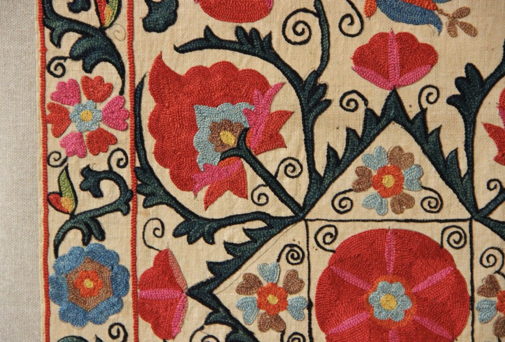 A rectangular mounted textile panel featuring intricate floral embroidery in red, blue, and green on a cream background with a decorative border. View V