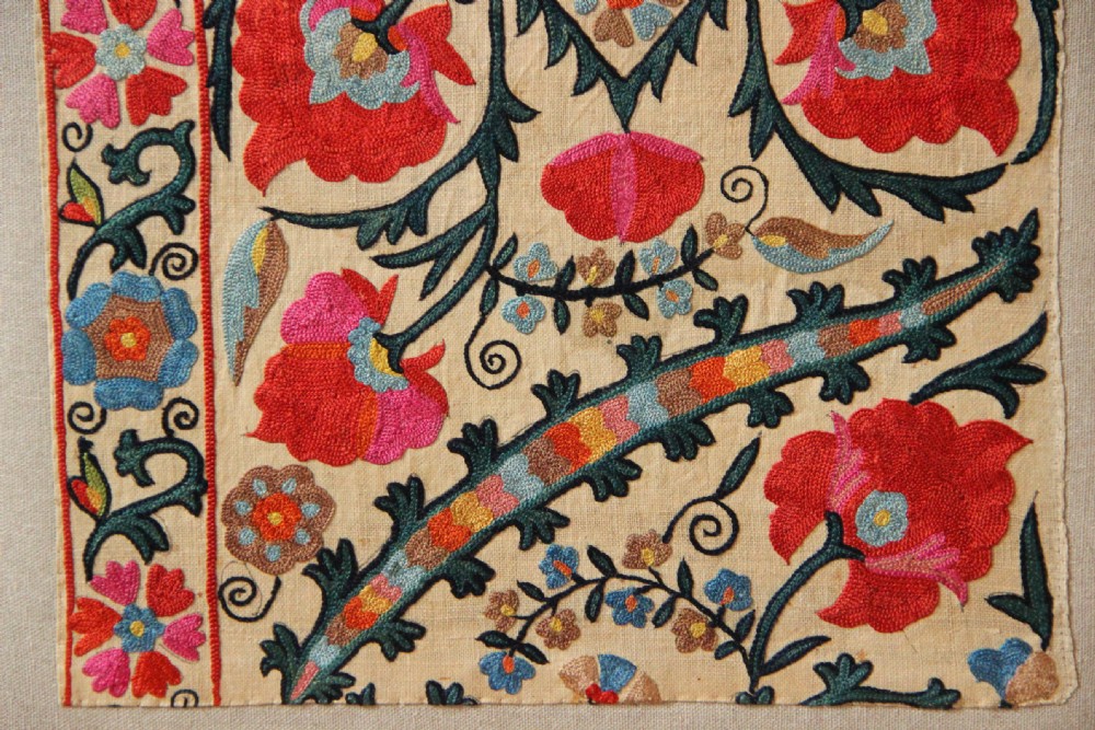 A rectangular mounted textile panel featuring intricate floral embroidery in red, blue, and green on a cream background with a decorative border. View VI