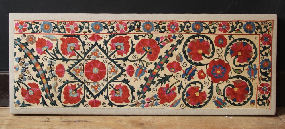 A rectangular mounted textile panel featuring intricate floral embroidery in red, blue, and green on a cream background with a decorative border. View VII