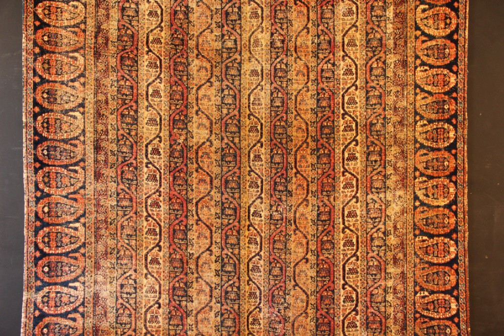 A large rectangular antique Persian Khorassan wool carpet featuring vertical striped pattern bands framed by a dark navy border filled with repeating boteh paisley motifs in rust and ochre tones. View II