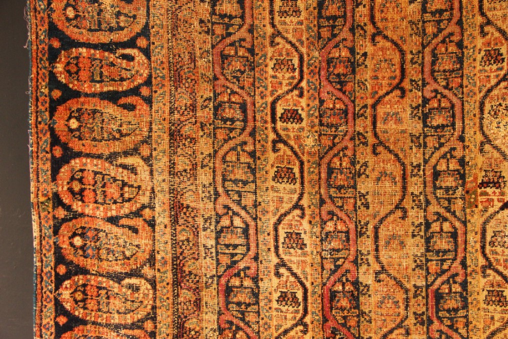 A large rectangular antique Persian Khorassan wool carpet featuring vertical striped pattern bands framed by a dark navy border filled with repeating boteh paisley motifs in rust and ochre tones. View VIII