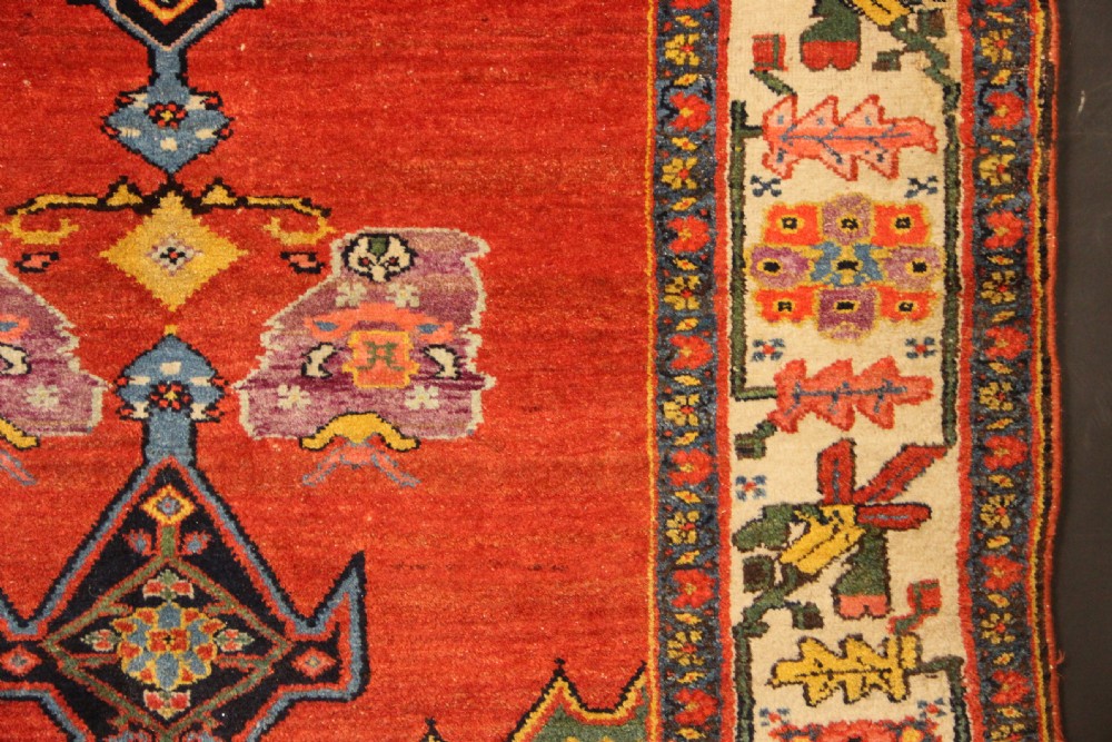 An antique long Bidjar carpet featuring a terracotta red field with central navy geometric medallions, green corner spandrels, and a cream floral border. View VI