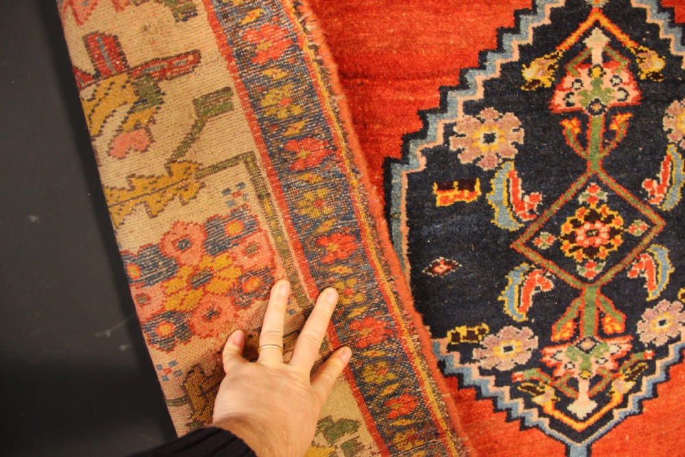 An antique long Bidjar carpet featuring a terracotta red field with central navy geometric medallions, green corner spandrels, and a cream floral border. View X
