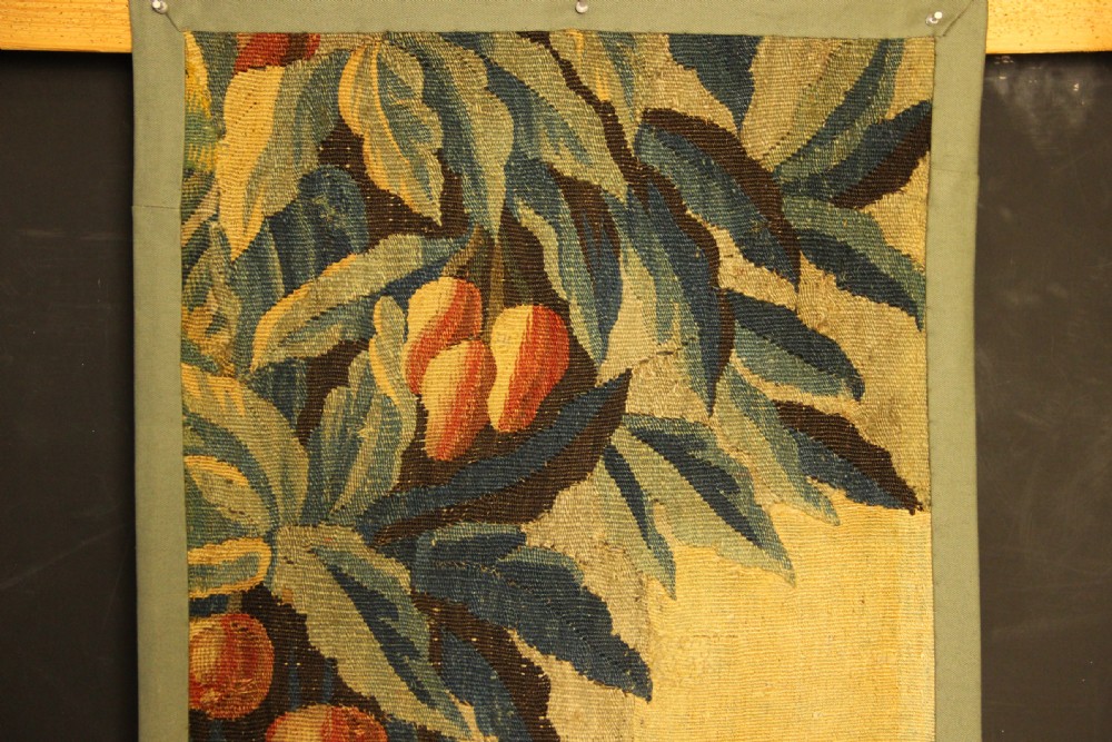 A vertical hand-woven textile panel depicting leafy foliage, fruit-bearing branches, small flowers, and a distant building in muted green, ochre, and blue tones. View III