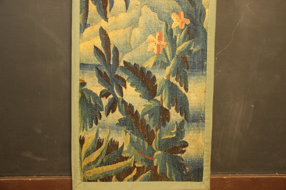 A vertical hand-woven textile panel depicting leafy foliage, fruit-bearing branches, small flowers, and a distant building in muted green, ochre, and blue tones. View V