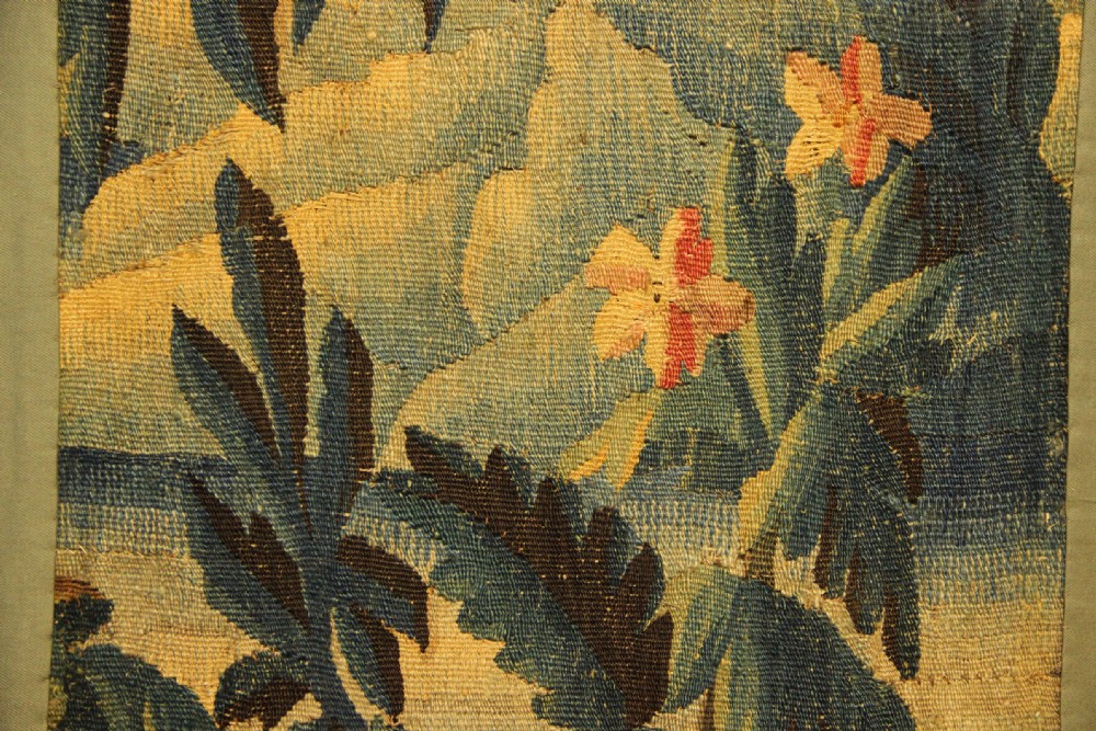 A vertical hand-woven textile panel depicting leafy foliage, fruit-bearing branches, small flowers, and a distant building in muted green, ochre, and blue tones. View VI