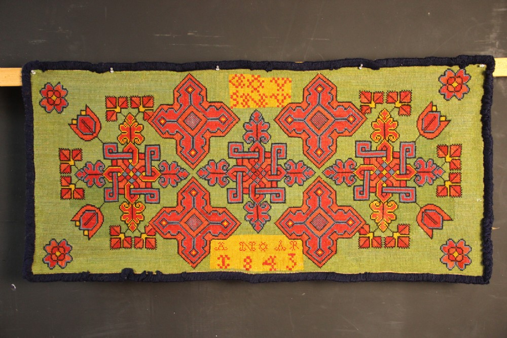 A rectangular 19th-century Swedish wool embroidery featuring geometric and floral motifs in red, orange, and blue against a green background, inscribed with the year 1843.