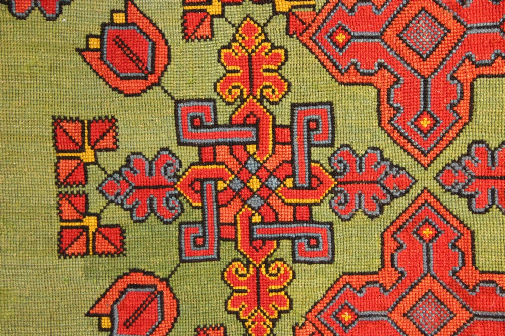 A rectangular 19th-century Swedish wool embroidery featuring geometric and floral motifs in red, orange, and blue against a green background, inscribed with the year 1843. View IV