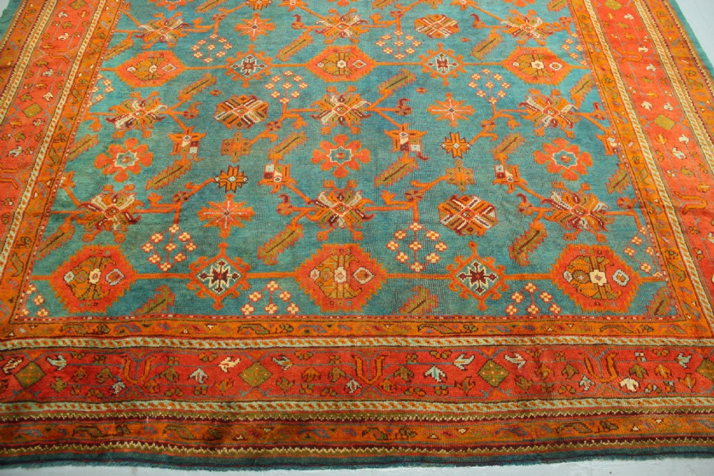 An antique Oushak wool rug displaying a teal field with geometric orange motifs and multi-layered terracotta borders. View X