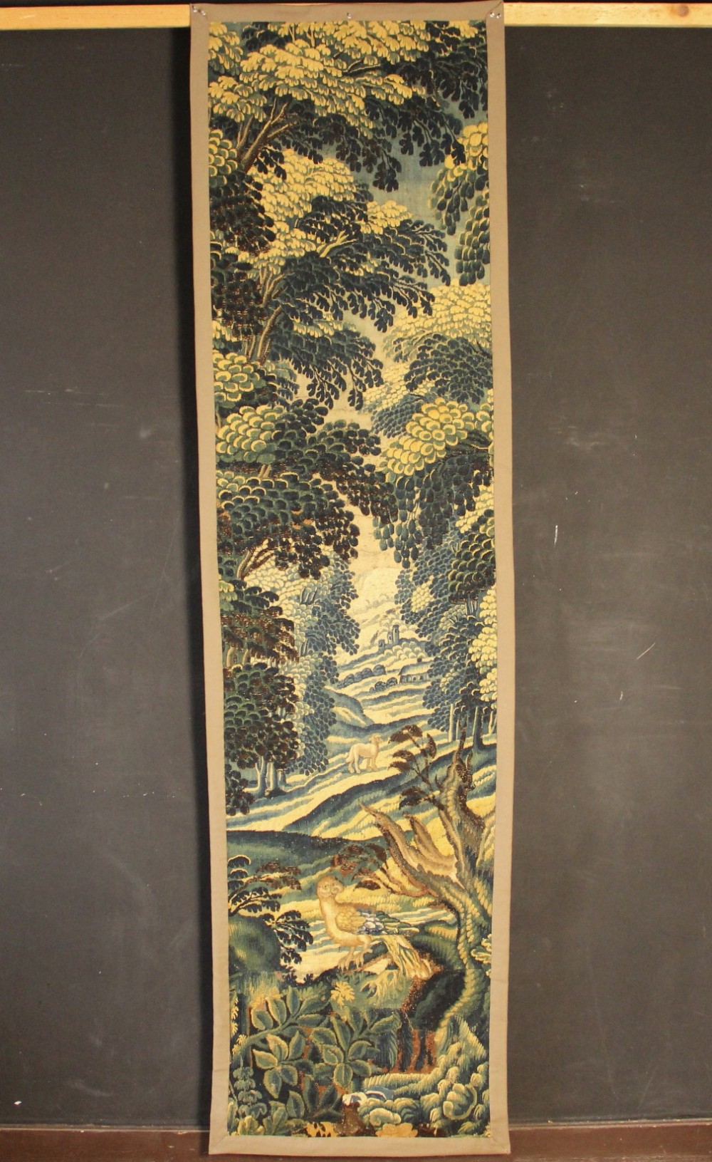 A narrow, vertical 17th-century Flemish verdure tapestry depicting a forest scene with lush foliage, trees, a bird, and distant rolling hills in shades of green and yellow.