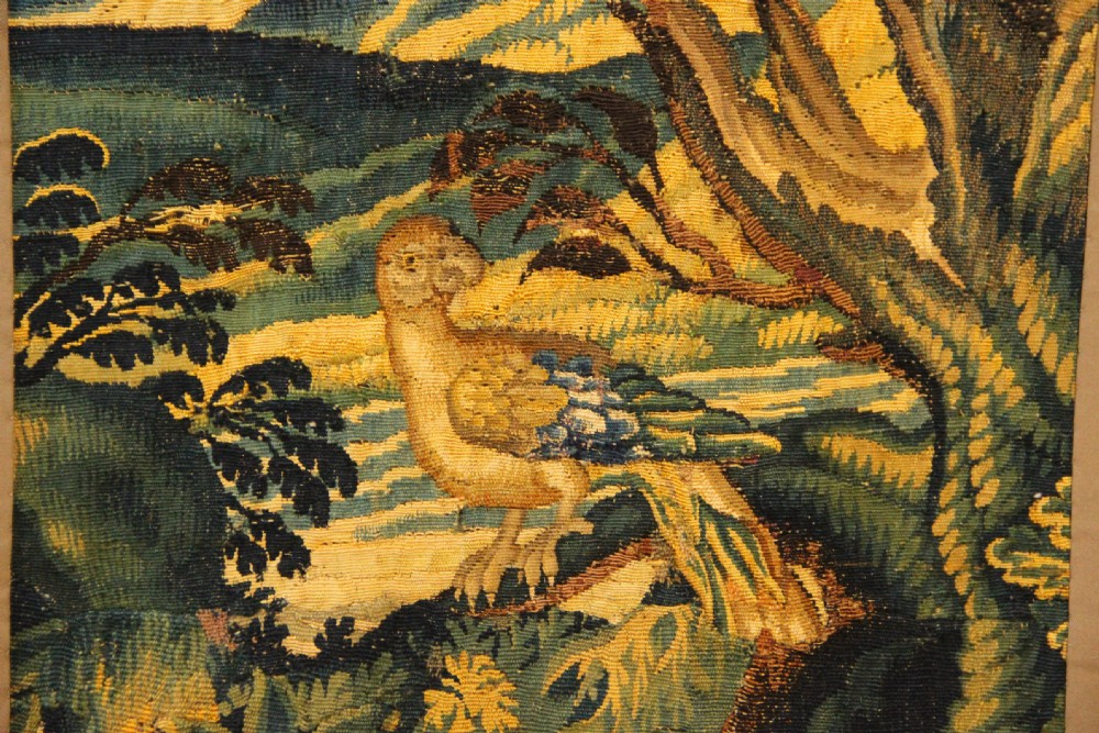 A narrow, vertical 17th-century Flemish verdure tapestry depicting a forest scene with lush foliage, trees, a bird, and distant rolling hills in shades of green and yellow. View V