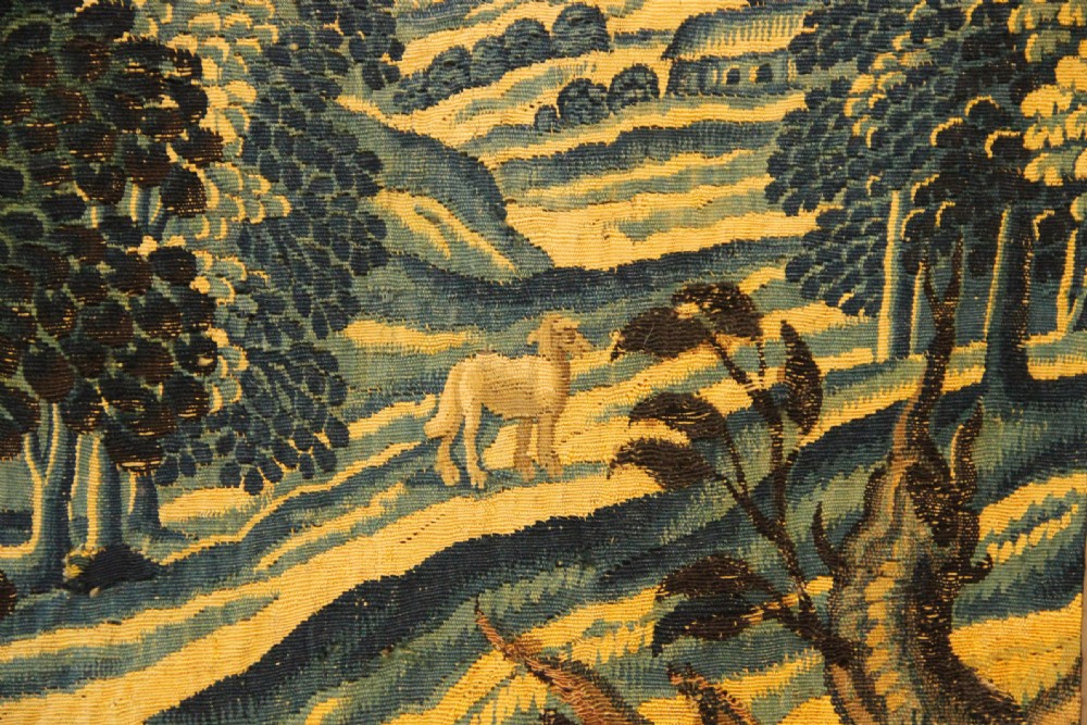 A narrow, vertical 17th-century Flemish verdure tapestry depicting a forest scene with lush foliage, trees, a bird, and distant rolling hills in shades of green and yellow. View VI