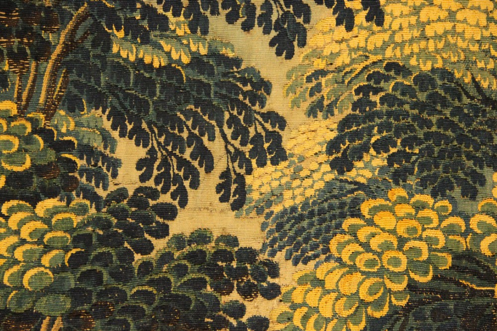 A narrow, vertical 17th-century Flemish verdure tapestry depicting a forest scene with lush foliage, trees, a bird, and distant rolling hills in shades of green and yellow. View VII