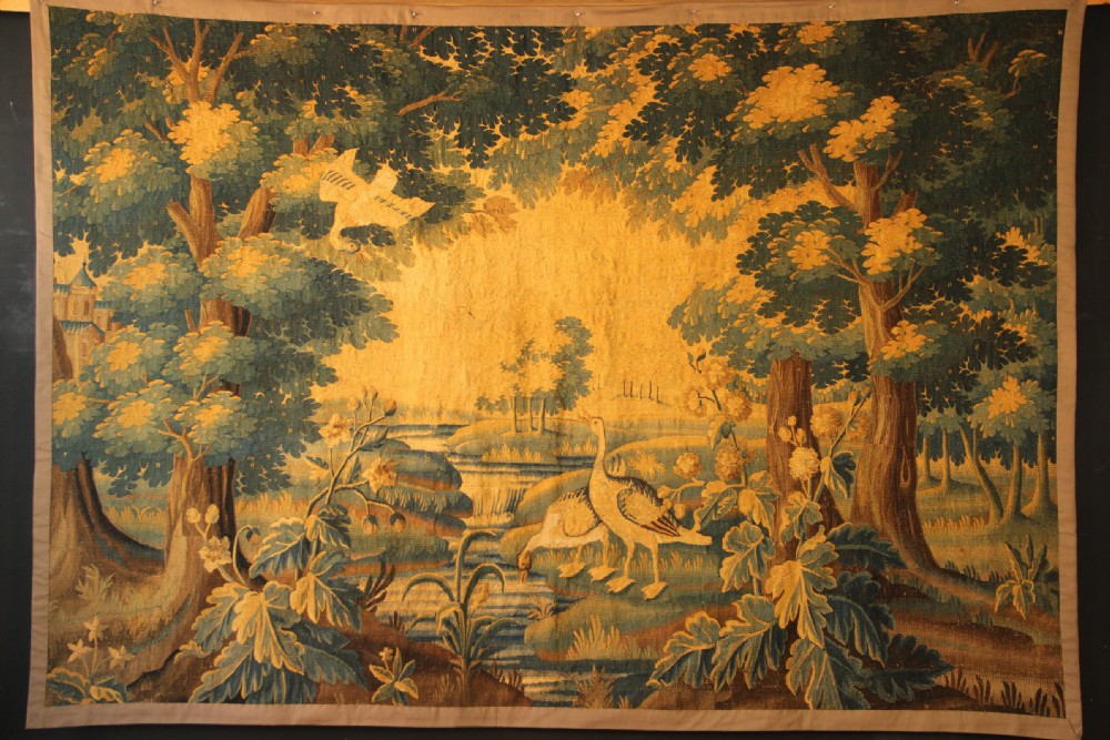A late 17th-century Flemish verdure wall tapestry depicting a woodland scene with tall trees, waterfowl near a stream, and distant architectural buildings in green and gold tones.