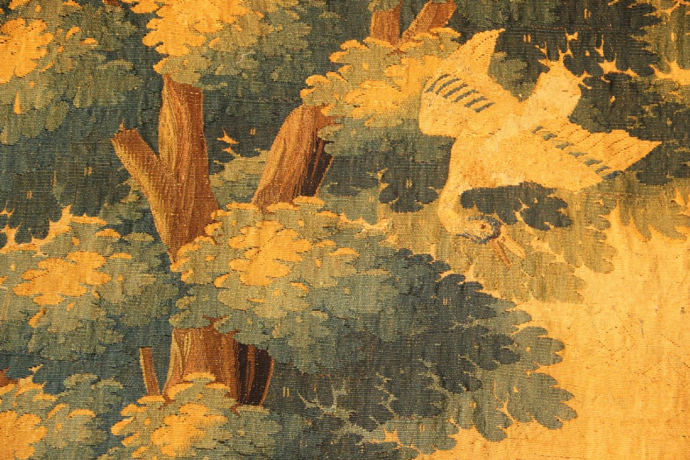 A late 17th-century Flemish verdure wall tapestry depicting a woodland scene with tall trees, waterfowl near a stream, and distant architectural buildings in green and gold tones. View II