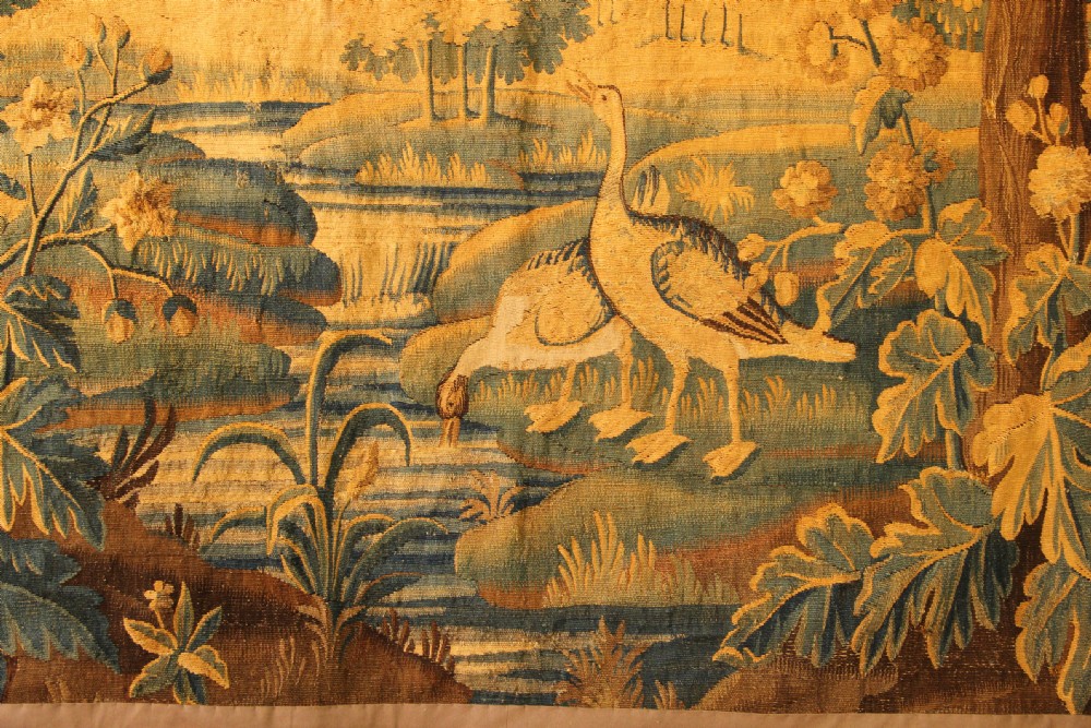 A late 17th-century Flemish verdure wall tapestry depicting a woodland scene with tall trees, waterfowl near a stream, and distant architectural buildings in green and gold tones. View III