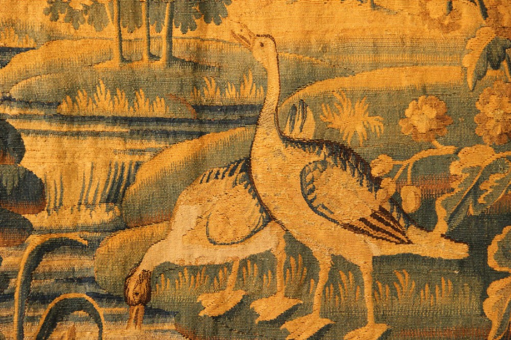 A late 17th-century Flemish verdure wall tapestry depicting a woodland scene with tall trees, waterfowl near a stream, and distant architectural buildings in green and gold tones. View V
