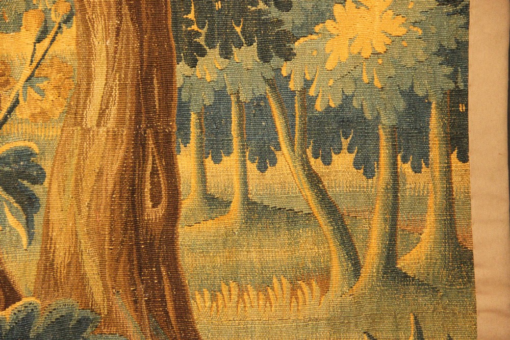 A late 17th-century Flemish verdure wall tapestry depicting a woodland scene with tall trees, waterfowl near a stream, and distant architectural buildings in green and gold tones. View VI