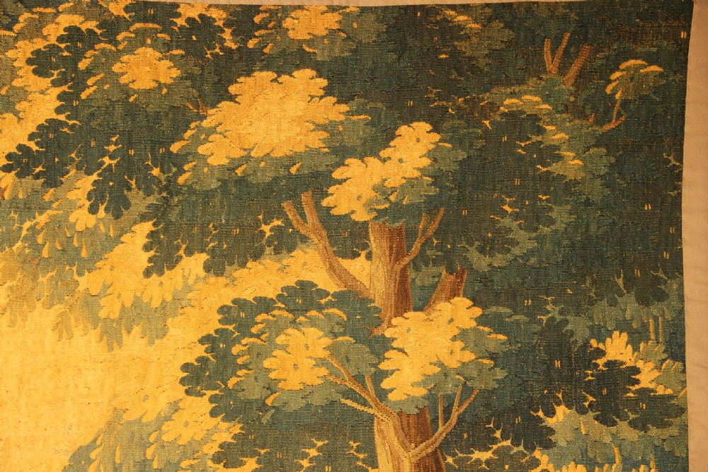 A late 17th-century Flemish verdure wall tapestry depicting a woodland scene with tall trees, waterfowl near a stream, and distant architectural buildings in green and gold tones. View VII