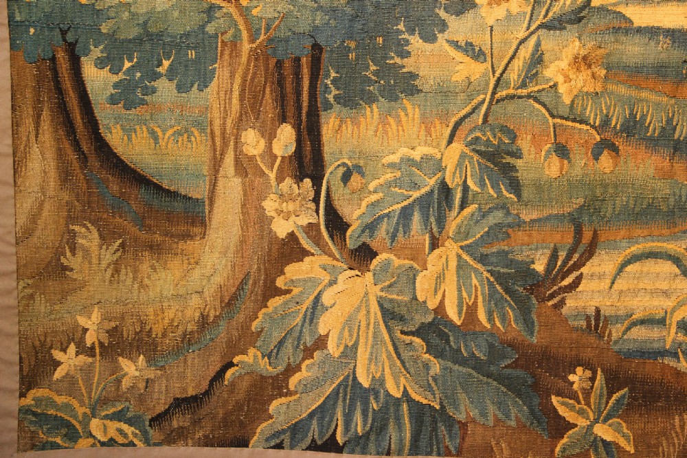 A late 17th-century Flemish verdure wall tapestry depicting a woodland scene with tall trees, waterfowl near a stream, and distant architectural buildings in green and gold tones. View VIII