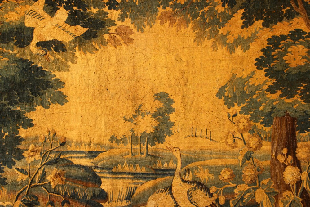 A late 17th-century Flemish verdure wall tapestry depicting a woodland scene with tall trees, waterfowl near a stream, and distant architectural buildings in green and gold tones. View IX