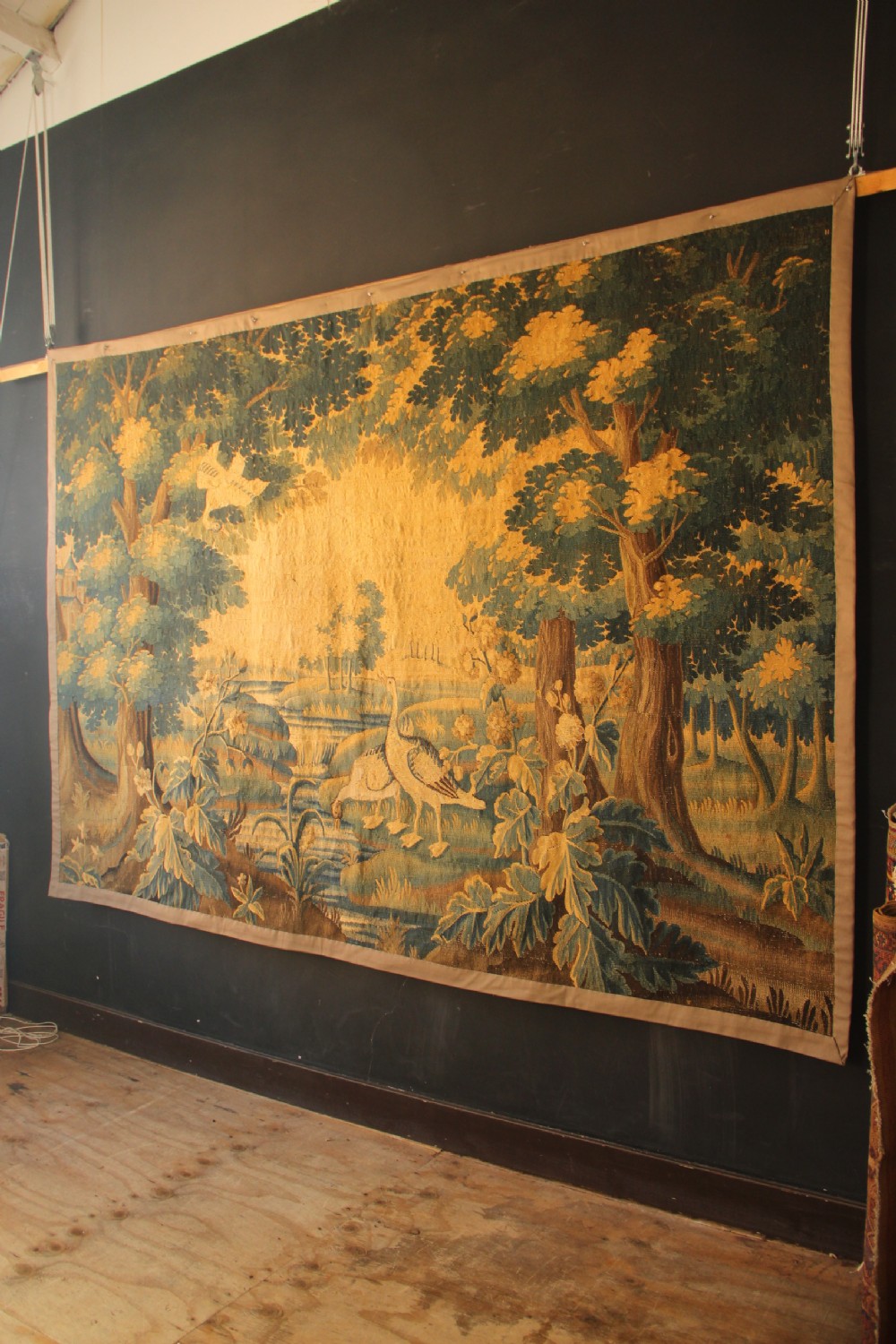 A late 17th-century Flemish verdure wall tapestry depicting a woodland scene with tall trees, waterfowl near a stream, and distant architectural buildings in green and gold tones. View X