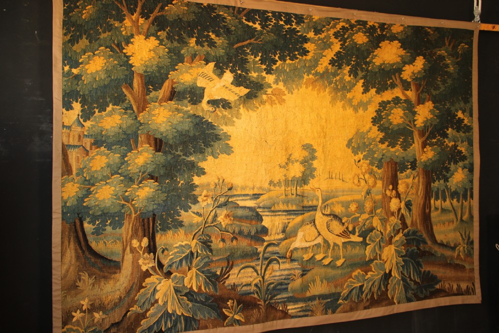 A late 17th-century Flemish verdure wall tapestry depicting a woodland scene with tall trees, waterfowl near a stream, and distant architectural buildings in green and gold tones. View XI