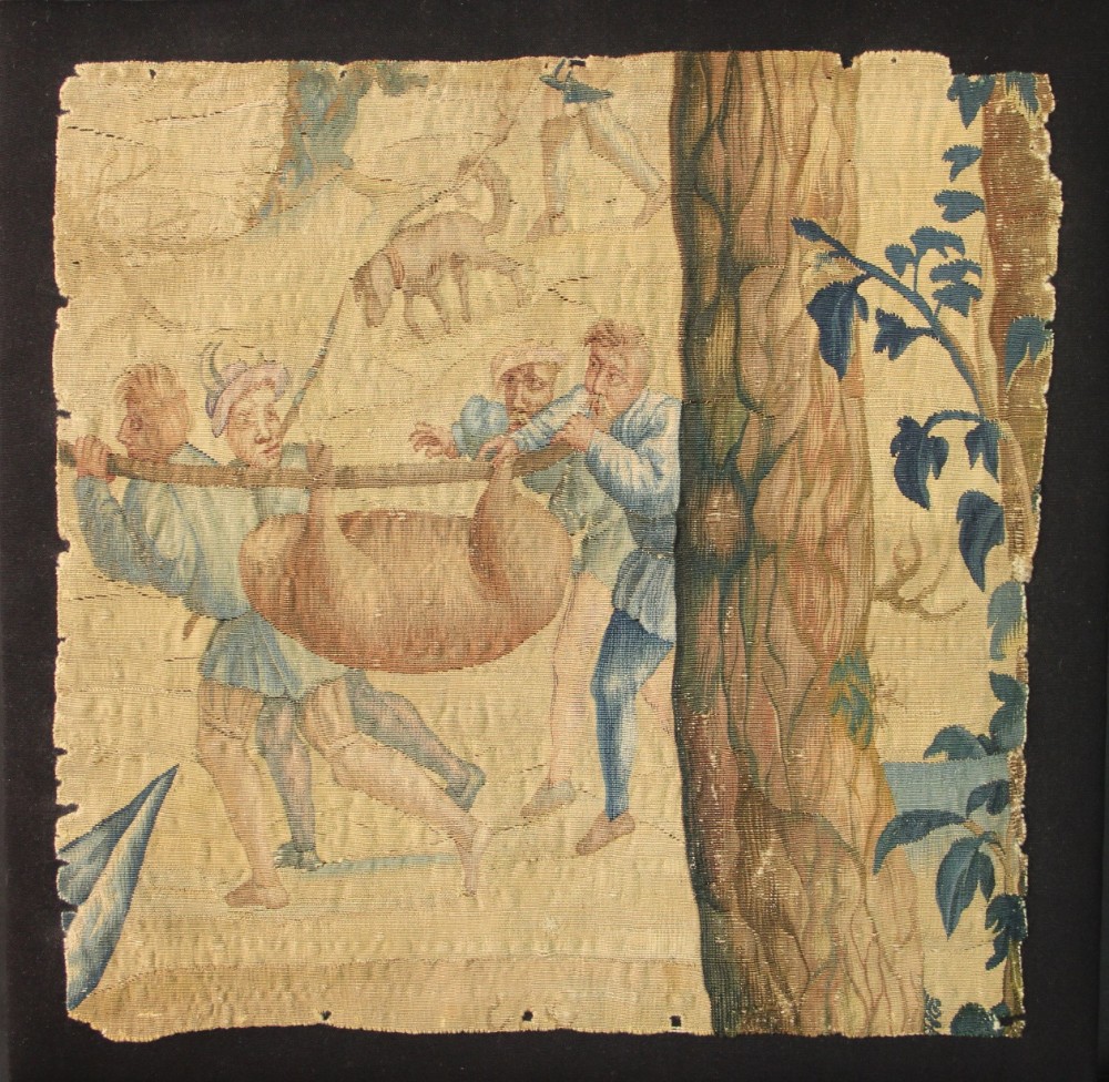 An early 17th-century square Flemish tapestry fragment depicting figures carrying a tied animal on a pole alongside a large tree trunk woven in beige, ochre, and blue tones.