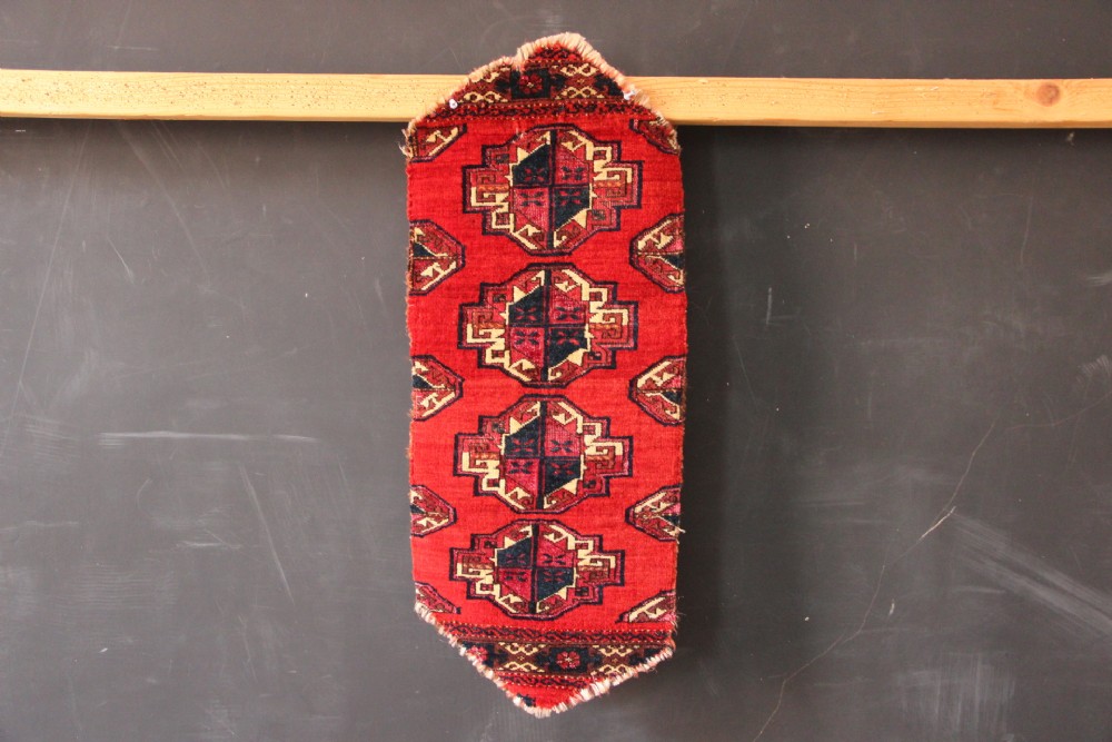 An antique vertical red wool textile fragment featuring repeating geometric Turkoman gol motifs in dark blue and cream.
