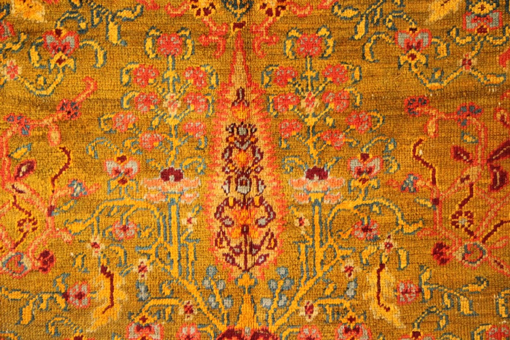 A large hand-knotted wool carpet featuring mustard yellow, olive green, and terracotta red tones with detailed floral and paisley motifs throughout. View III