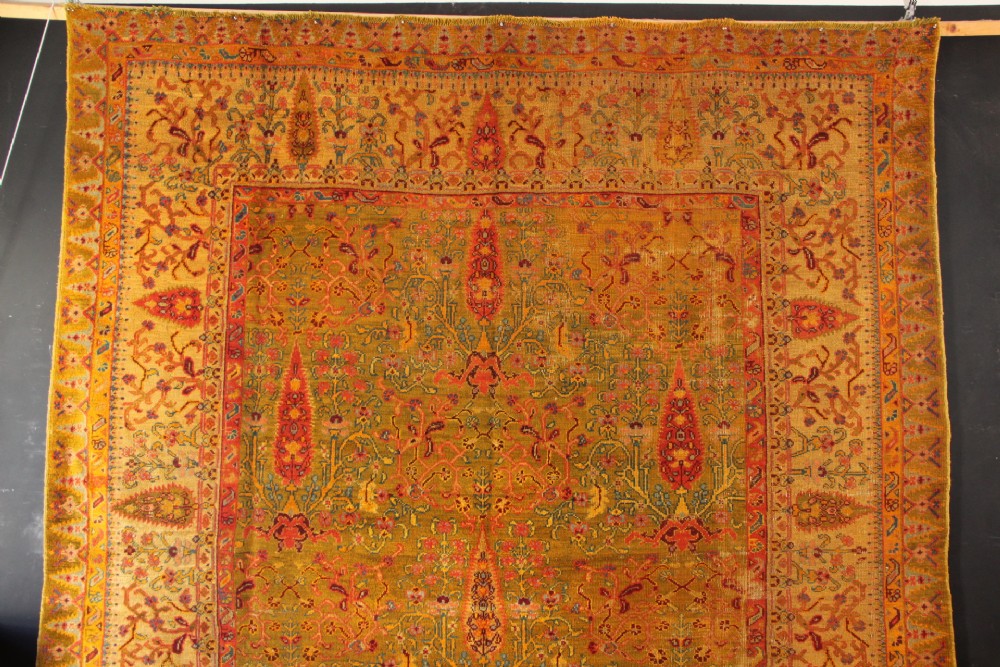 A large hand-knotted wool carpet featuring mustard yellow, olive green, and terracotta red tones with detailed floral and paisley motifs throughout. View X