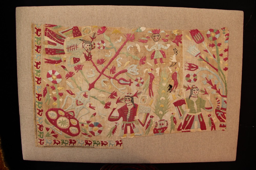 An antique hand-embroidered linen textile fragment from Skyros featuring red and green silk threads depicting human figures, birds, and floral motifs.