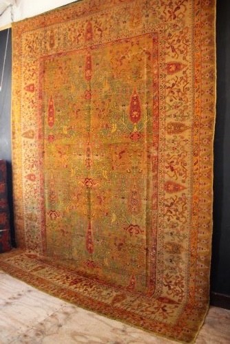 A large hand-knotted wool carpet featuring mustard yellow, olive green, and terracotta red tones with detailed floral and paisley motifs throughout. View VII