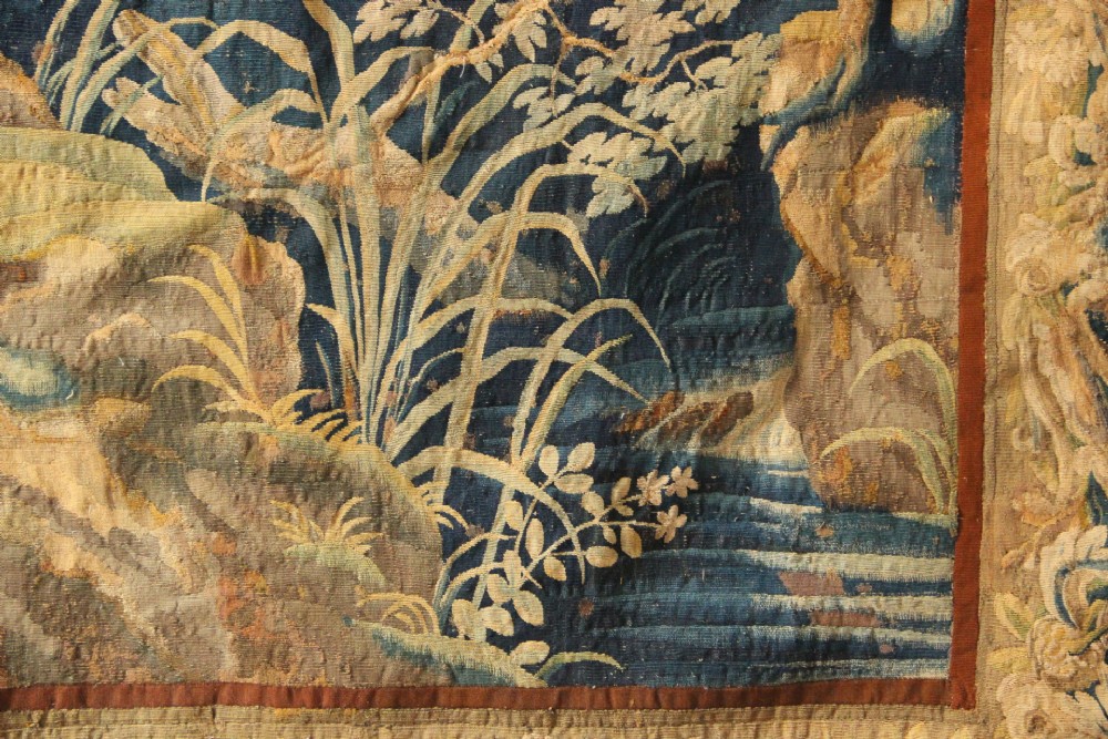 A large woven Flemish verdure tapestry featuring a dense forest landscape with trees, botanical plants, a central chateau view, and a decorative foliage border. View III