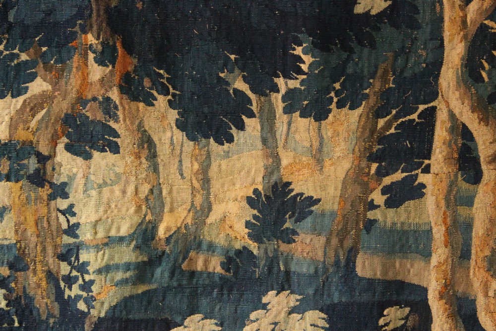 A large woven Flemish verdure tapestry featuring a dense forest landscape with trees, botanical plants, a central chateau view, and a decorative foliage border. View IV