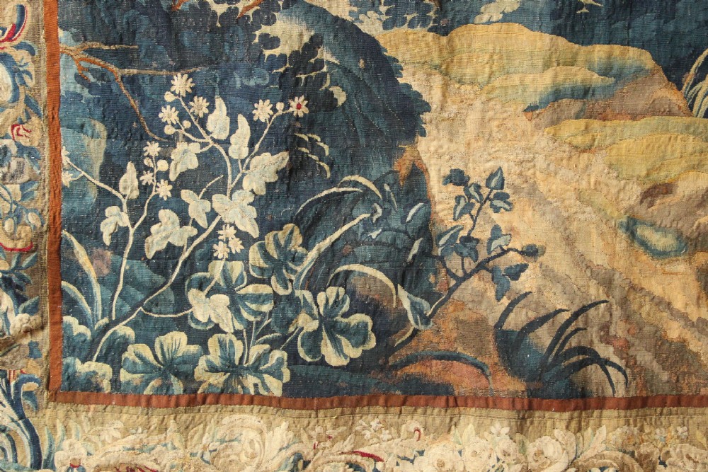 A large woven Flemish verdure tapestry featuring a dense forest landscape with trees, botanical plants, a central chateau view, and a decorative foliage border. View V