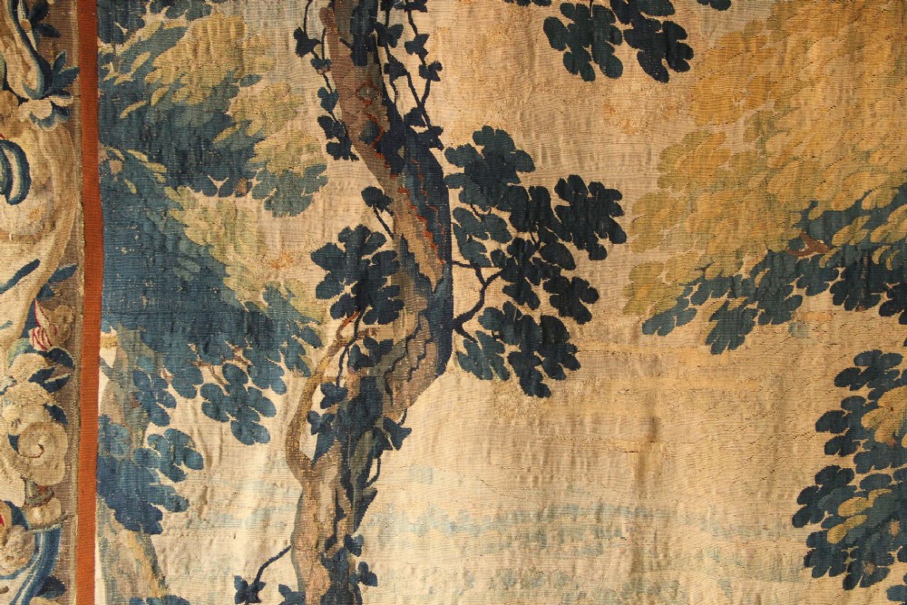 A large woven Flemish verdure tapestry featuring a dense forest landscape with trees, botanical plants, a central chateau view, and a decorative foliage border. View VI