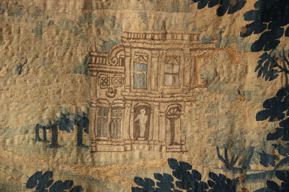 A large woven Flemish verdure tapestry featuring a dense forest landscape with trees, botanical plants, a central chateau view, and a decorative foliage border. View VII