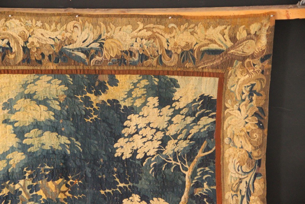 A large woven Flemish verdure tapestry featuring a dense forest landscape with trees, botanical plants, a central chateau view, and a decorative foliage border. View VIII