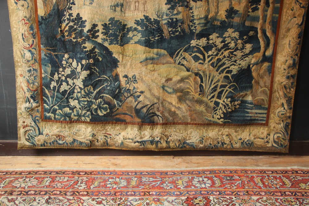 A large woven Flemish verdure tapestry featuring a dense forest landscape with trees, botanical plants, a central chateau view, and a decorative foliage border. View IX